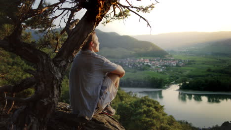 male-looking-at-the-sunset-under-a-tree,-mountain-landscape-background,-golden-hour-river-danube-valley,-overview,-looking-out,-peaceful,-detox,-digital-detox,-outdoor-nature,-detox