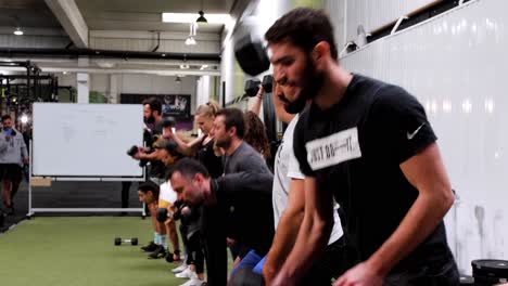 several-athletes-lined-up-to-take-part-in-a-crossfit-session,-all-stooping-down-at-the-same-time-to-pick-up-an-alter-on-the-floor-with-one-hand-and-lift-it