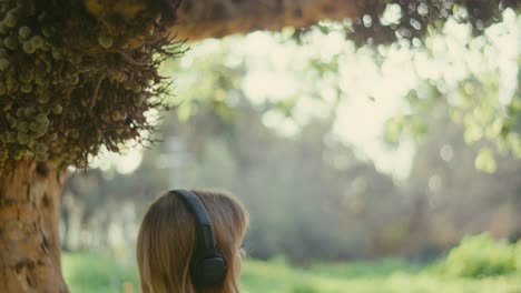 Lady-with-loose-hair-sits-on-bench-under-tree-listening-to-music-on-headphones