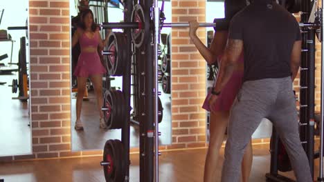 Hispanic-girl-squats-at-the-gym-with-help-from-a-trainer