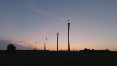 Aerial-establishing-view-wind-turbines-generating-renewable-energy-in-a-wind-farm,-evening-after-the-sunset-golden-hour,-countryside-landscape,-high-contrast-silhouettes,-drone-dolly-shot-moving-right
