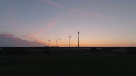 Aerial-establishing-view-wind-turbines-generating-renewable-energy-in-a-wind-farm,-evening-after-the-sunset-golden-hour,-countryside-landscape,-high-contrast-silhouettes,-drone-shot-moving-forward