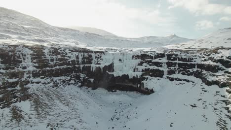 4K-Aerial-drone-shot-of-140-meter-tall-frozen-waterfall-Fossurin-I-Fossá,-the-largest-waterfall-on-the-Faroe-Islands-on-Streymoy-on-a-cold-and-sunny-winter-day-in-a-landscape-covered-in-snow
