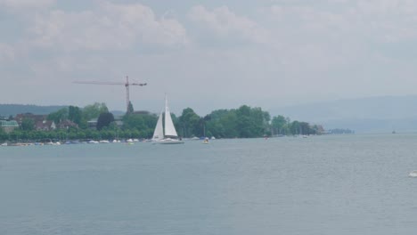 Scenic-drone-footage-of-a-sailing-boat-with-a-Swiss-flag-sailing-on-a-beautiful-lake-at-the-foot-of-the-Alps,-not-far-from-a-village-located-on-the-coast