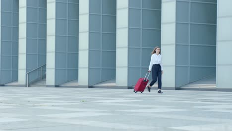 Skinny-girl-in-elegant-clothes,-walking-with-red-suitcase-in-front-of-modern-building