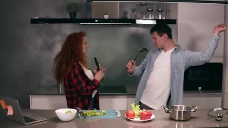 Happy-carefree-young-family-couple-husband-and-wife-singing-in-soup-ladle-and-pasta-tongs-microphone-together-prepare-healthy-food-romantic-dinner-breakfast-cooking-dancing-having-fun-listen-music-on-kitchen