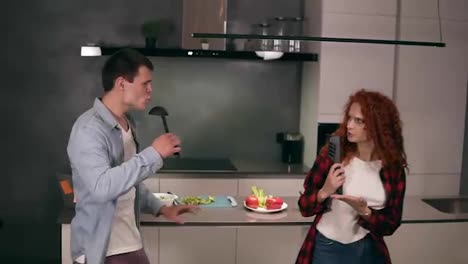 Happy-carefree-young-family-couple-husband-and-wife-singing-in-soup-ladle-and-pasta-tongs-microphone-together-prepare-healthy-food-romantic-dinner-breakfast-cooking-dancing-having-fun-listen-music-on-kitchen.-Slow-motion