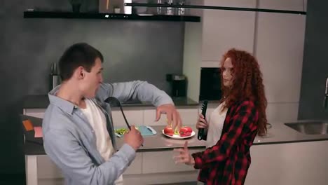 Caucasian-young-family-couple-husband-and-redheaded-wife-singing-in-soup-ladle-and-pasta-tongs-microphone-together-prepare-using-kitchen-as-a-stage,-dancing-having-fun-listen-music-on-kitchen.-Slow-motion