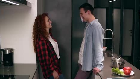 Young-happy-couple-or-friends-talking-in-the-kitchen-and-looking-at-each-other.-Long-haired-curly-woman-in-plaid-shirt-standing-in-front-her-boyfriend-in-blue-shirt-in-modern-loft-designed-kitchen