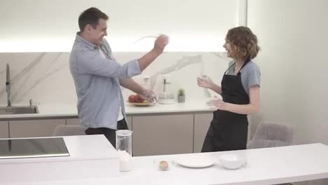 Cute-positive-girl-blowing-flour-on-boyfriend's-face-in-the-kitchen,-man-is-throwing-back-to-the-face.-Happy-young-couple-fooling-around-while-cooking-food.-Side-view,-slow-motion