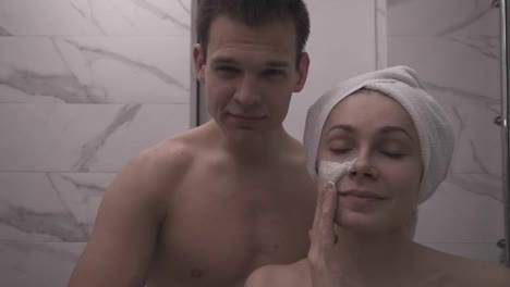 Happy-positive-young-couple-man-and-woman-having-fun-while-standing-in-a-bathroom-after-shower.-Both-playfully-applying-white-mask-on-face.-Having-fun.-Portrait.-Slow-motion