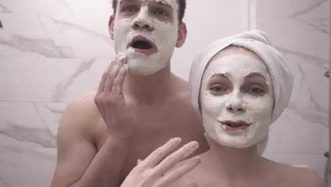 Happy-positive-young-couple-man-and-woman-having-fun-while-standing-in-a-bathroom-after-shower.-Both-playfully-applying-white-mask-on-face.-Having-fun,-girl-explaining-mask-properties-to-her-boyfriend