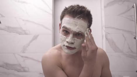 Portrait-of-young-handsome-man-is-applying-fresh-white-thick-mask-and-scrub-on-the-face.-Natural-organic-cosmetics-for-skin-care.-Man-after-shower,-face-skin-care
