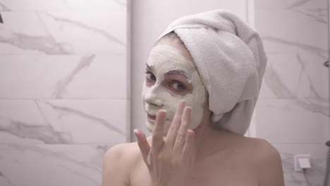 Portrait-of-young-pretty-woman-with-white-towel-on-head-is-applying-fresh-white-thick-mask-and-scrub-on-the-face.-Natural-organic-cosmetics-for-skin-care.-Woman-after-shower,-face-skin-care