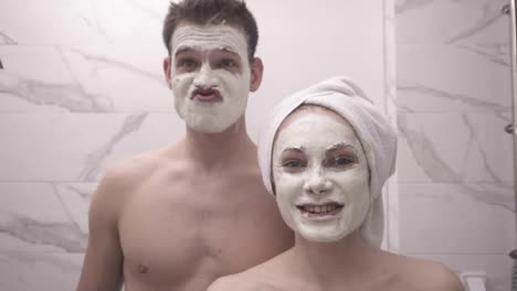 Crazy,-funny-couple-standing-in-front-mirror-in-bathroom-after-shower---man-topless,-girl-with-white-towel-on-head---making-funny-faces,-posing,-grimasing-with-white-mask-on-face