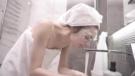 A-woman-standing-in-the-bathroom-near-the-sink-in-white-towel-on-head-and-body-washes-off-a-white-cosmetic-mask-with-water-from-the-tap.-Side-view