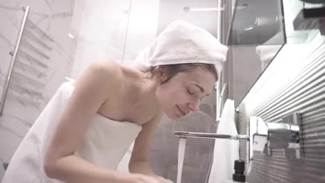 A-woman-standing-in-the-bathroom-near-the-sink-in-white-towel-on-head-and-body-washes-her-face-with-water-from-the-tap.-Looking-to-the-mirror-reflection-and-smiling.-Low-angle-view
