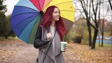Young-woman-with-red-hair-walking-in-autumn-park-and-drinking-coffee-from-a-paper-cup-while-holding-colorful-umbrella.-Girl-in-warm-coat-enjoying-cool-fall-weather-with-a-cup-of-hot-drink