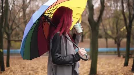 Side-view:-young-woman-with-red-hair-walking-in-autumn-park-and-drinking-coffee-from-a-paper-cup-while-holding-colorful-umbrella.-Girl-in-warm-coat-enjoying-cool-fall-weather-with-a-cup-of-hot-drink