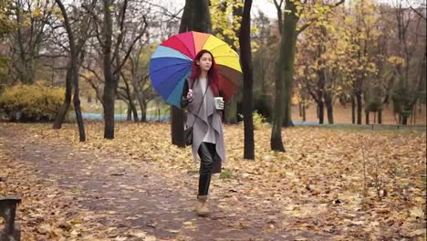 Young-stylish-woman-with-red-hair-walking-in-autumn-park-and-drinking-coffee-from-a-paper-cup-while-holding-colorful-umbrella.-Girl-in-warm-coat-enjoying-cool-fall-weather-with-a-cup-of-hot-drink
