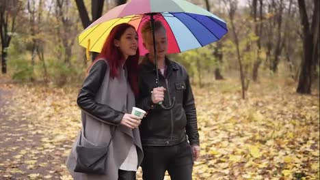 Young-happy-couple-walking-together-in-autumn-park-holding-a-colorful-umbrella.-Attractive-woman-with-red-hair-is-holding-a-paper-cup-with-coffee.-They-are-talking-and-holding-hands