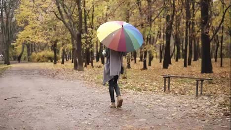 Back-view-of-a-young-woman-with-red-hair-walking-in-autumn-park-with-colorful-rainbow-umbrella-and-holding-paper-cup-with-coffee.-Girl-in-warm-coat-enjoying-cool-fall-weather-with-a-cup-of-hot-drink