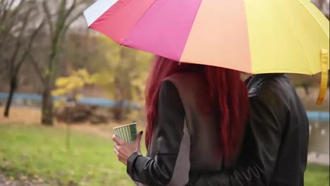 Happy-couple-walking-together-in-autumn-park-holding-a-colorful-umbrella.-Attractive-man-in-leather-jacket-is-embracing-his-girlfriend.-Beautiful-woman-with-red-hair-is-holding-a-paper-cup-with-coffee