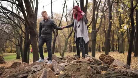 Romantic-young-couple-walking-on-the-rocks-in-autumn-park-during-the-day-holding-hands.-Caring-boyfriend-is-holsing-his-girlfriend's-hand.-Warm-weather-in-autumn
