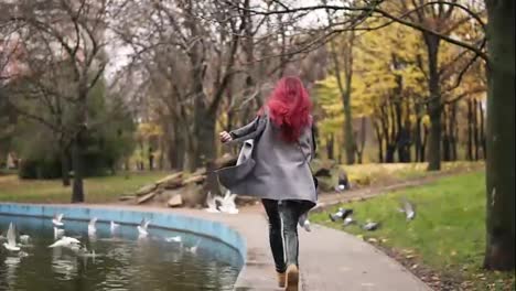 Back-view-of-attractive-woman-with-red-hair-running-towards-pigeons-and-making-them-fly-away-in-a-park-with-artificial-lake