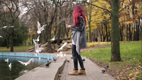 Side-view-of-red-haired-woman-in-warm-coat-feeding-the-gulls-by-the-pond-in-park-in-autumn.-Slowmotion-shot