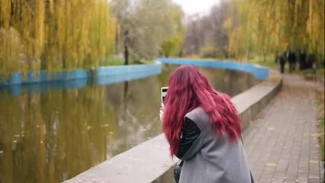 Junge-Attraktive-Frau-Mit-Rotem-Haar,-Die-Mit-Ihrem-Smartphone-Ein-Foto-Macht,-Während-Sie-An-Einem-Künstlichen-Teich-In-Einem-Herbstpark-Steht