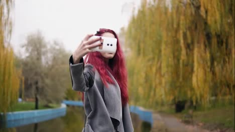 Young-attractive-woman-with-red-hait-making-selfie-on-her-smartphone-while-standing-by-an-artificial-pond-in-an-autumn-park