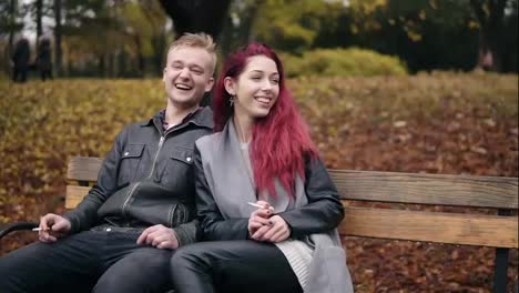 Young-attractive-couple-sitting-on-a-bench-in-autumn-park-and-talking.-Attractive-woman-with-red-hair-is-smoking