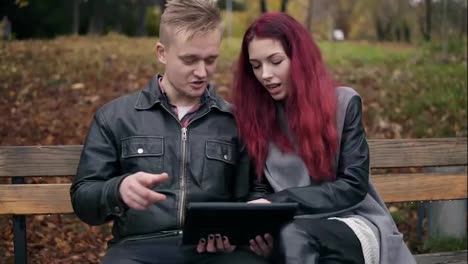 Young-woman-with-red-hair-and-attractive-man-in-a-leather-jacket-sitting-on-a-bench-in-park-and-using-a-digital-talbe-while-discussing-someting.-They-are-choosing-a-trip-and-shopping-online