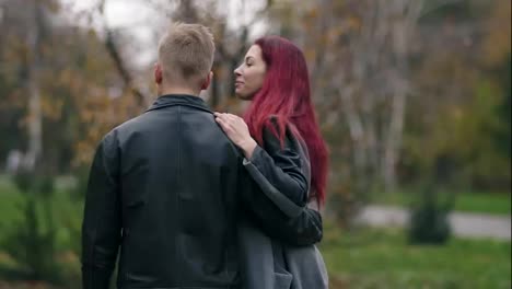 Romantic-young-couple-walking-in-autumn-park-during-the-day.-Back-view-of-young-blonde-man-in-leather-jacket-embracing-his-girlfriend.-Loving-couple-talking-and-spending-time-together