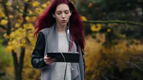 Young-businesswoman-with-red-hair-walks-in-the-autumn-park-and-communicates-via-tablet-pc.-Young-attractive-woman-with-digital-tablet-computer-in-hands-communicates-using-earphones