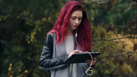 Young-beautiful-woman-with-red-hair-walks-in-the-autumn-park-and-listens-to-music-via-tablet-pc.-Young-attractive-woman-with-digital-tablet-computer-in-hands