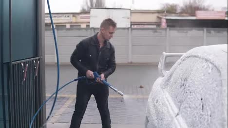 Young-man-in-leather-jacket-applying-foam-on-silver-sportcar-with-special-jet-on-self-service-carwash