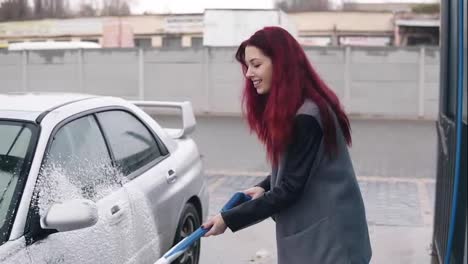 Young-woman-with-red-hair-applying-foam-on-her-silver-sportcar-with-special-jet-on-self-service-carwash