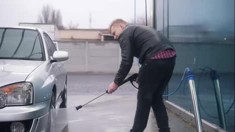 Young-attractive-stylish-man-washing-his-silver-sportcar-with-water-jet-on-self-service-carwash.-He-is-carefully-cleaning-the-tyres-of-the-car