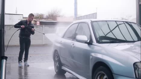 Young-attractive-stylish-man-carefully-washing-his-silver-sportcar-with-water-jet-on-self-service-carwash.-Slowmotion-shot