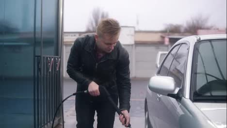Young-attractive-stylish-man-washing-his-silver-sportcar-with-water-jet-on-self-service-carwash.-He-is-carefully-cleaning-the-tyres-of-the-car.-Slowmotion-shot
