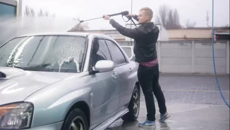 Young-attractive-stylish-man-is-carefully-washing-his-silver-sportcar-with-water-jet-on-self-service-carwash.-Slowmotion-shot