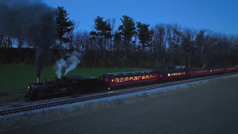 Drone-Slightly-Ahead-and-Parallel-Night-View-of-a-Steam-Passenger-Train-Stopped-in-Farmlands-Blowing-Lots-of-Smoke-Seeing-the-Lights-in-the-Coaches
