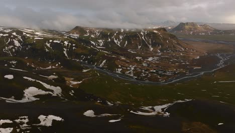 Luftpanorama-Landschaftsansicht-Eines-Flusses,-Der-An-Einem-Bewölkten-Und-Nebligen-Tag-Durch-Berge-Fließt,-Die-Mit-Schmelzendem-Schnee-Bedeckt-Sind,-In-Island