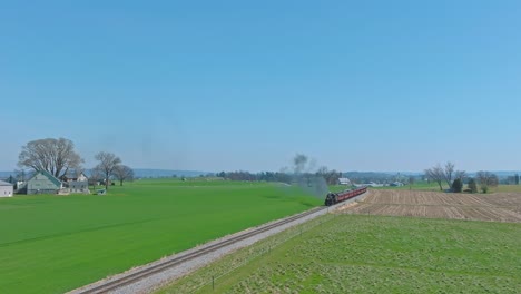 A-Drone-Stationary-View-of-a-Steam-Passenger-Train-Approaching-Thru-Farmlands,-Blowing-Smoke-on-a-Spring-Day
