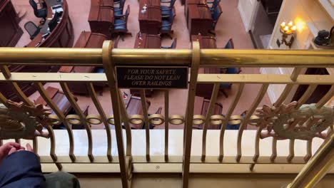 Looking-at-a-sign-that-says,-for-your-safety-please-do-not-lean-on-railing-with-person-or-kid-sitting-in-front-of-a-brass-railing-that-over-looks-Senate-Chamber-in-Utah-State-Capital