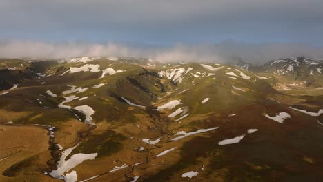 Vista-Aérea-Del-Paisaje-De-Un-Río-Que-Fluye-A-Través-De-Un-Valle-Montañoso,-Cubierto-De-Nieve-Derretida,-En-Islandia