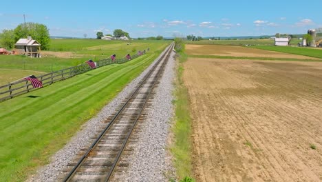 A-Drone-Static-View-of-a-Single-Empty-Rail-Road-Track-That-Goes-Thru-Green-Farmlands-With-a-Fence-With-American-Flags-on-it,-on-a-Spring-Day