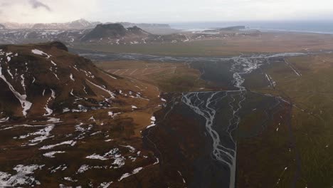 Aerial-landscape-view-of-a-river-flowing-through-mountains-covered-in-melting-snow,-on-a-cloudy-and-foggy-day,-in-Iceland